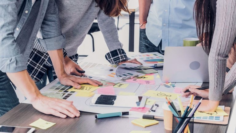 four people standing around a desk. no faces are visible. they are sticking post it notes to papers on a desk