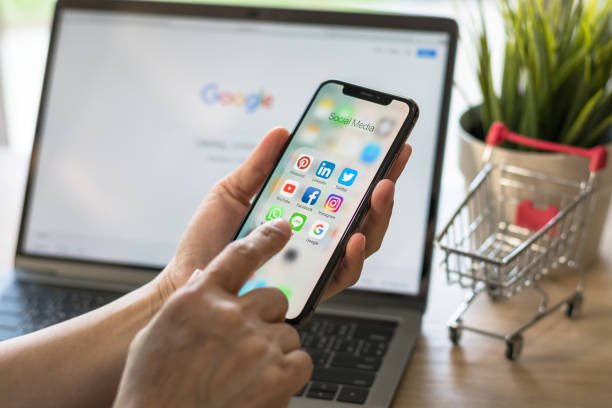 Person holds a smartphone in one hand and points to apps with one finger. There is a laptop in the background with google website on display. there is a tiny shopping cart in the background