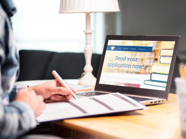 Person writing on a form with laptop open screen says "send your application"